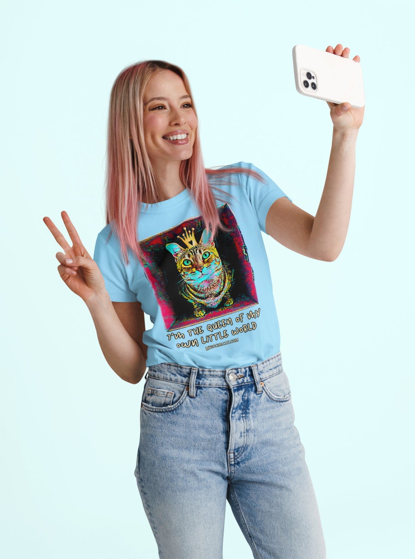 young woman with blond long hair standing against light blue background holding her phone smiling and making a peace sign with her other hand, wearing light blue jeans and a light blue tshirt with a graphic print of a cat sitting in the box looking at camera with a crown on its head that says, i'm the queen of my own little world mysocialcat.com