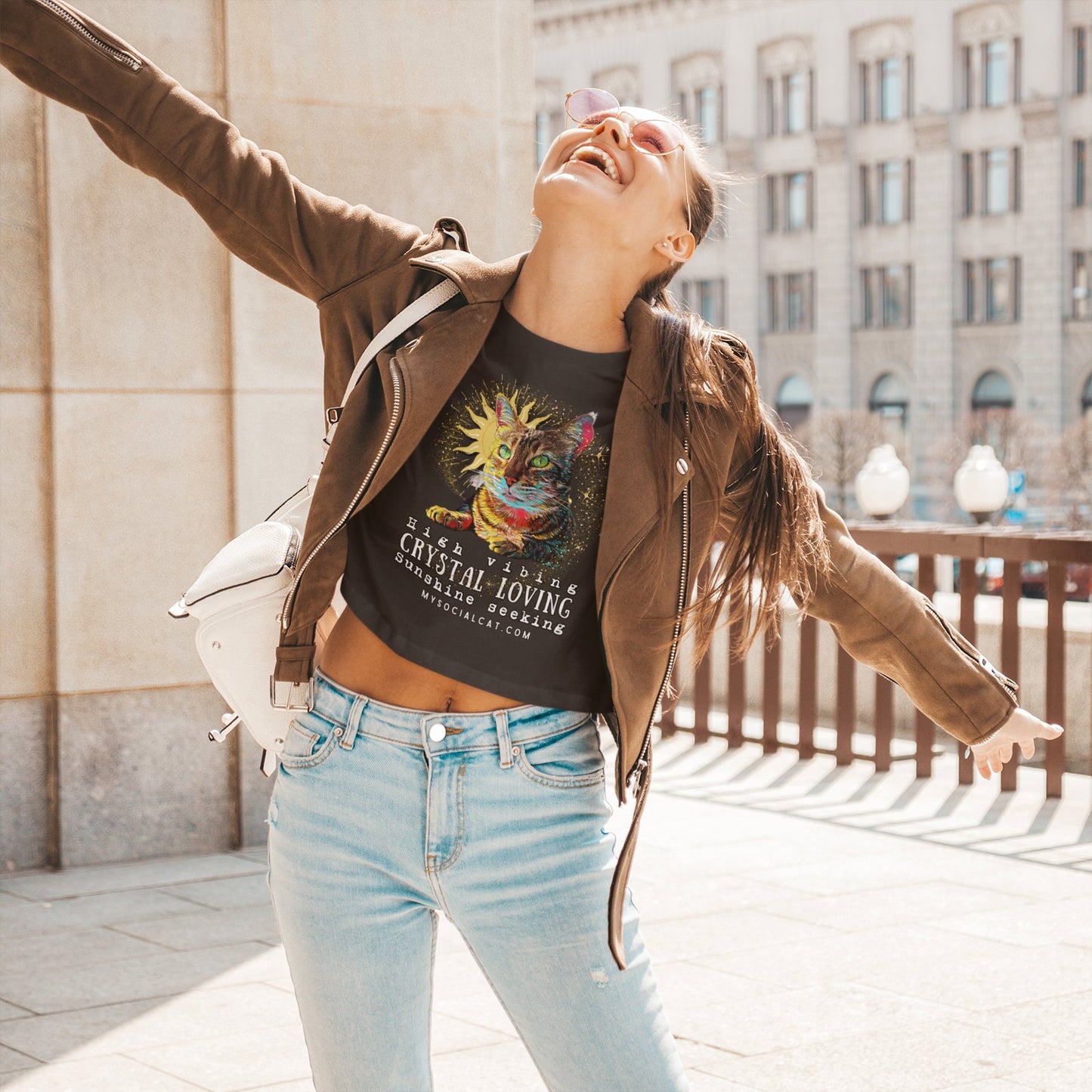 young woman outside in the city with arms out smiling to the sun wearing a brown leather jacket, light jeans and a black crop top with a cat face that says, high vibing crystal loving sunshine seeking mysocialcat.com 