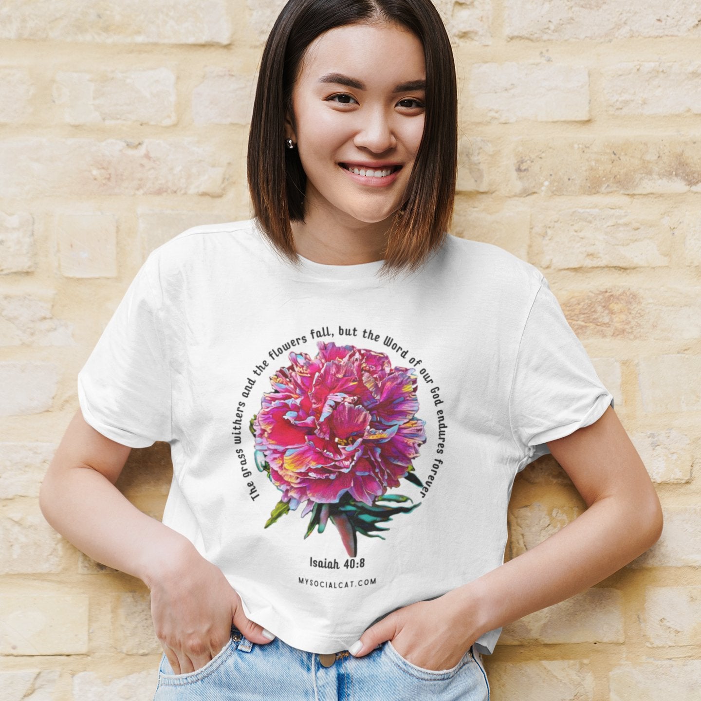 beautiful young Asian woman standing outside against a tan brick wall, with her hands in jeans pockets, smiling, wearing a white crop top featuring a pink peony and words across and around, grass withers, flowers fade but the word of our god endures forever mysocialcat.com 