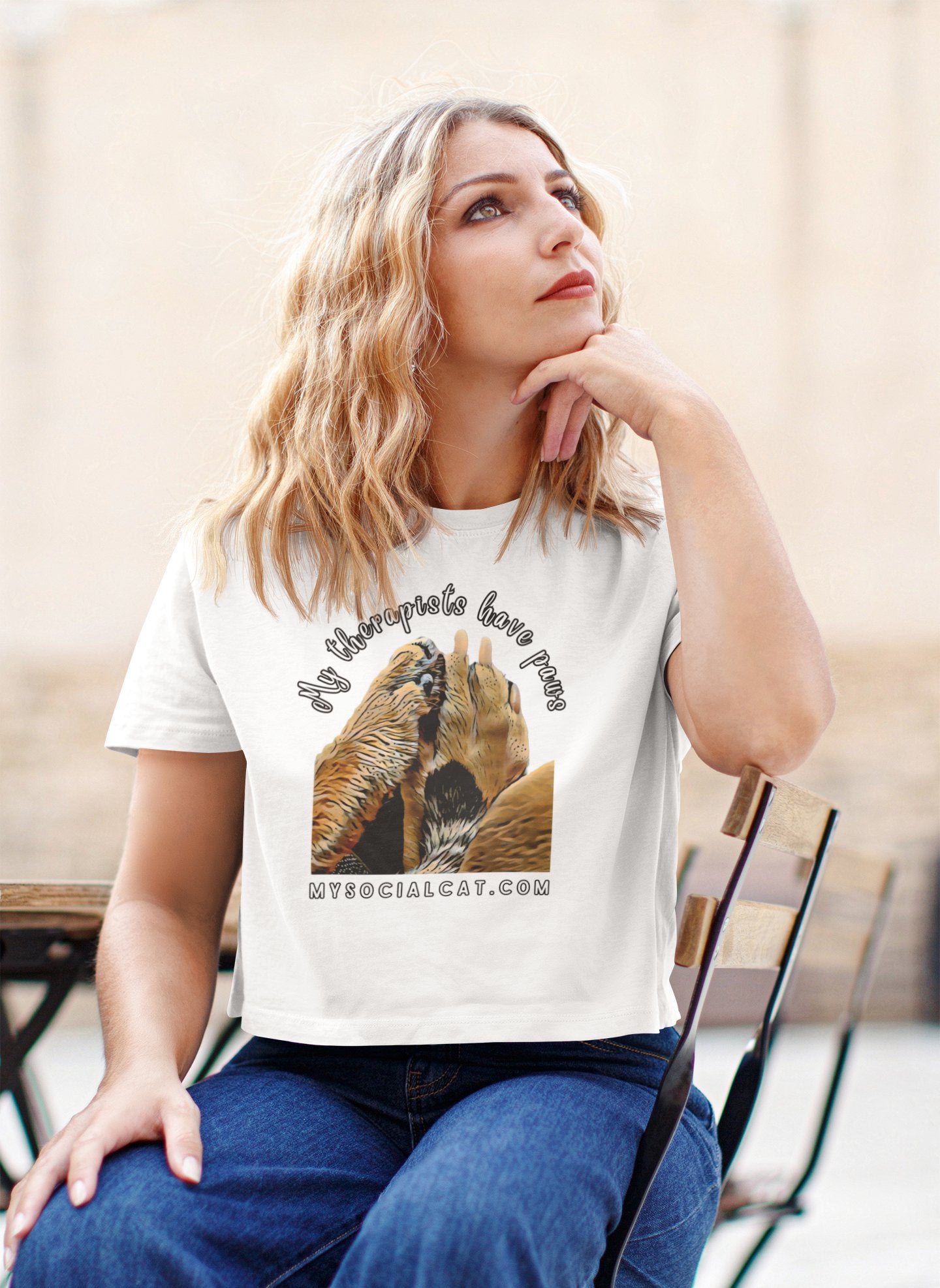 a blond wavy hair woman sitting in a chair with her hand under her chin looking up wearing blue jeans and a white crop top featuring two paws, cat and dog, that says, my therapists have paws, mysocialcat.com 