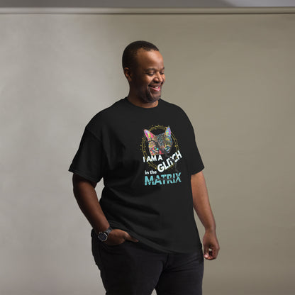 A man wearing a black graphic t-shirt with a colorful cat illustration and the text 'I AM A GLITCH IN THE MATRIX' in white and teal, standing against a neutral gray wall.