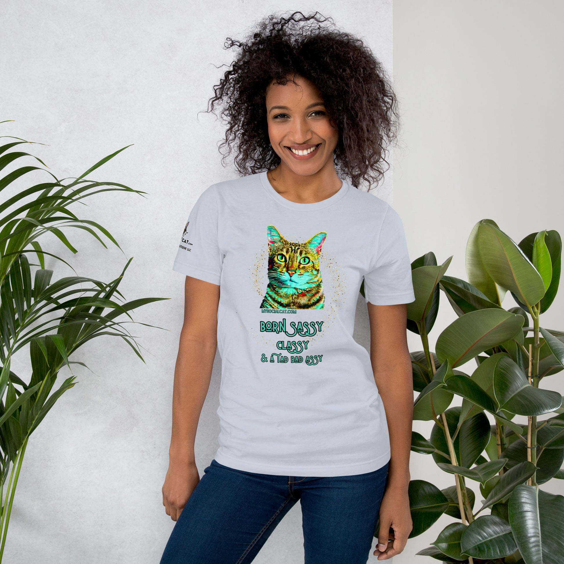 young woman standing against white background with houseplants wearing light blue tshirt smiling